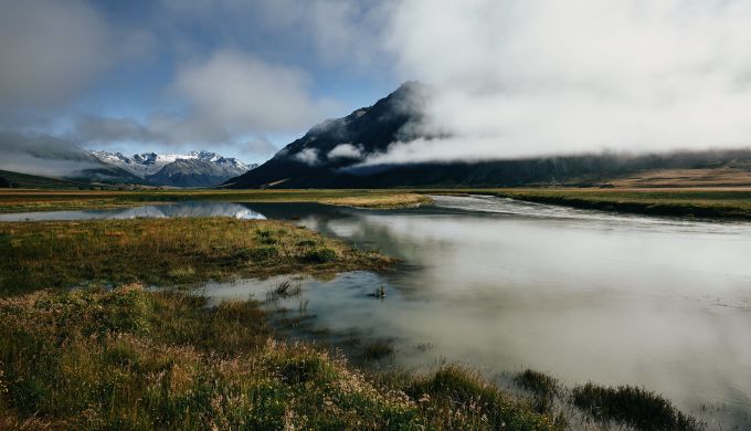 A valley view with distant mountains and low cloud reflected in a small lagoon.