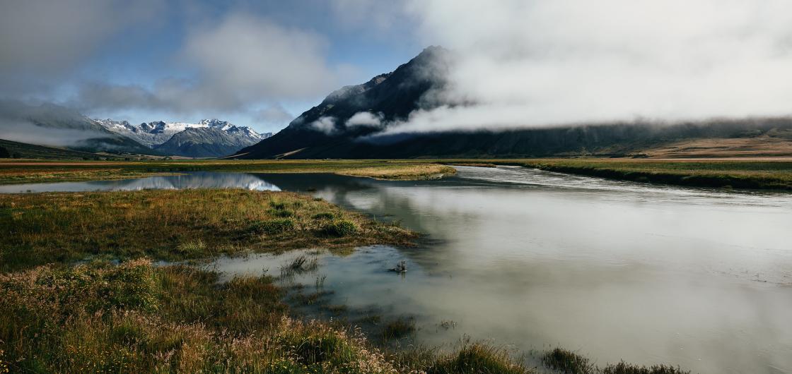A valley view with distant mountains and low cloud reflected in a small lagoon.