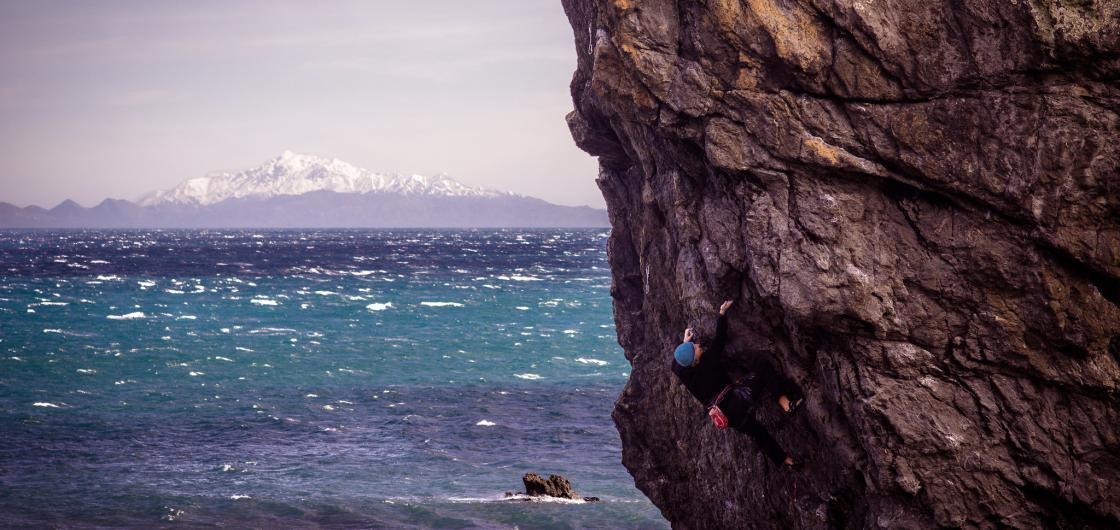 Man climbs steep rock with sea and mountain in background